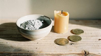 Керамическая миска с пеплом, догоревшая свеча и потёртые монеты на деревянной полке в утреннем свете