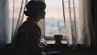 Женщина сидит у окна на рассвете, чашка чая остывает рядом, спокойный задумчивый свет